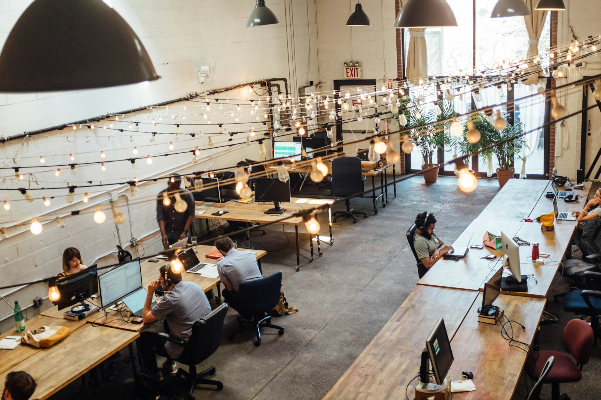 Multiple people working at long wooden desks under industrial pendant lights.